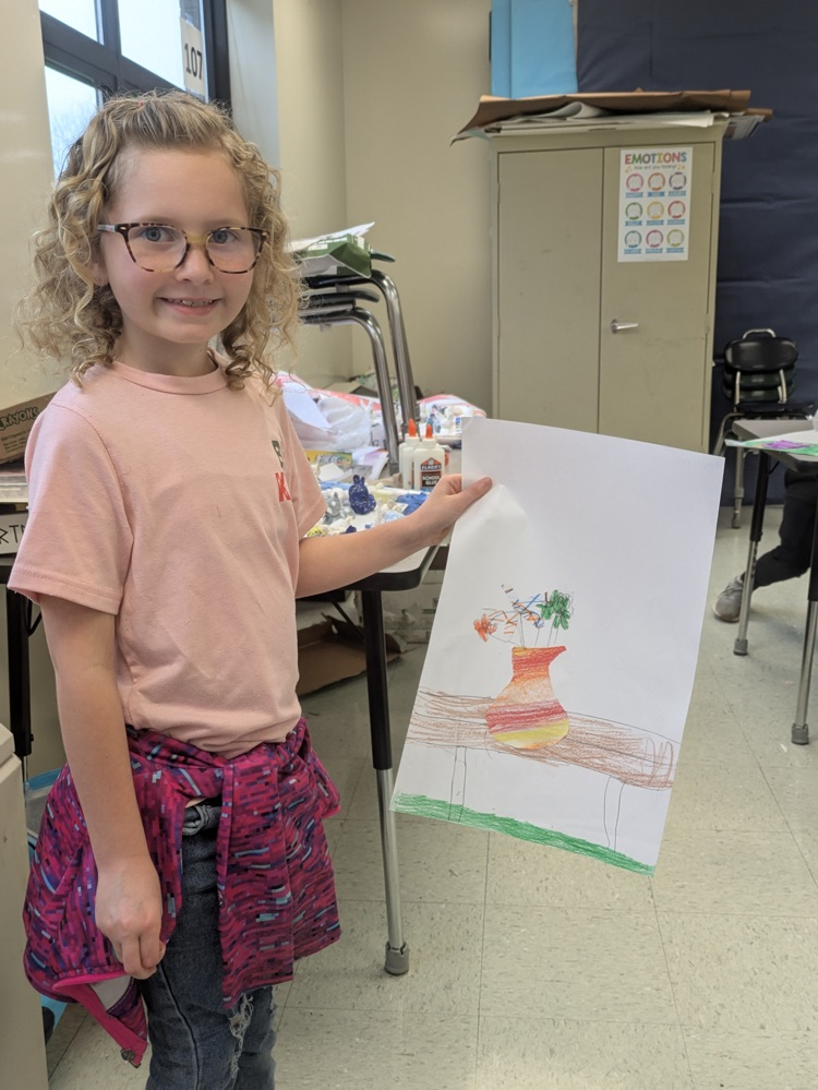 A girl smiles and holds up an art piece of a vase of flowers on a table.