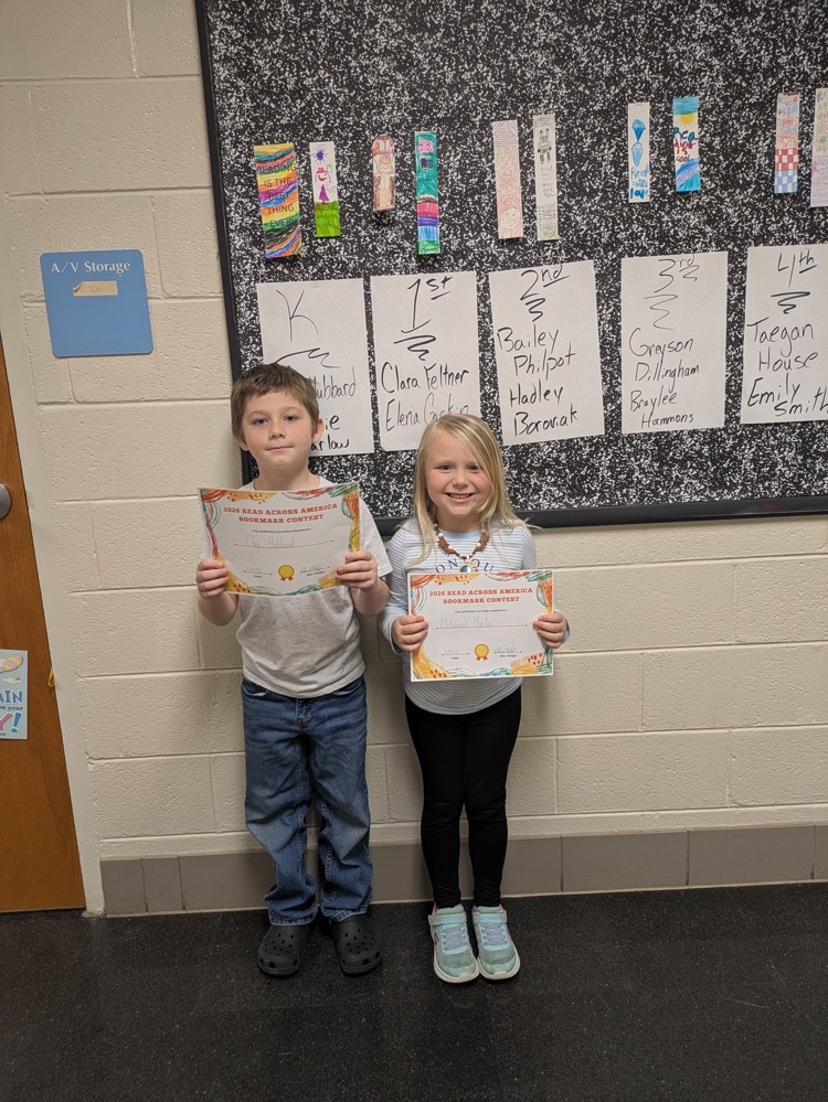 A boy and girl hold up their award certificates while standing in front of a bulletin board decorated with the winning bookmarks  