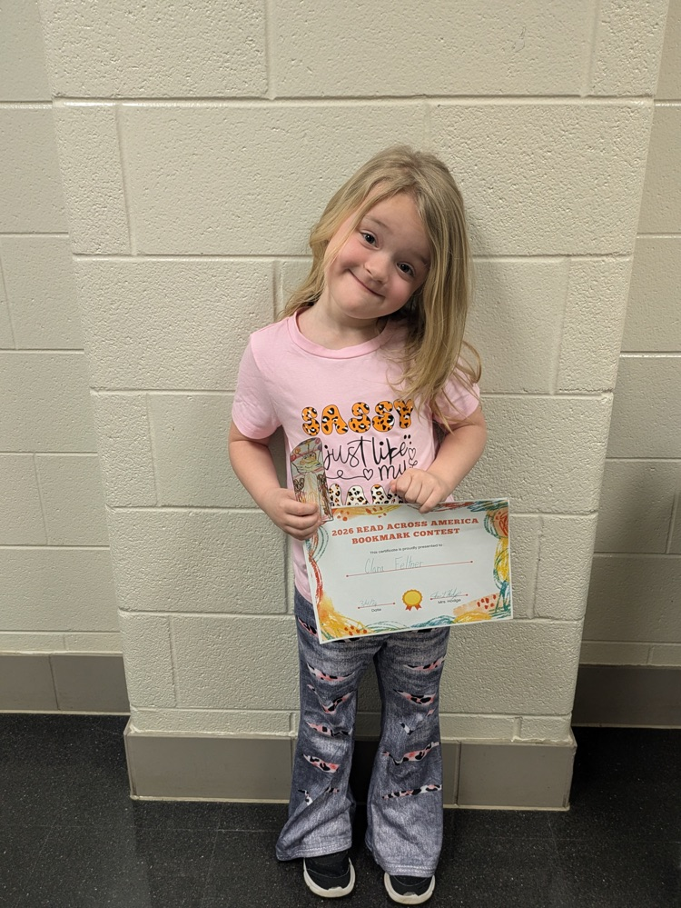 A girl holds her award certificate while standing in front of a bulletin board decorated with the winning bookmarks  