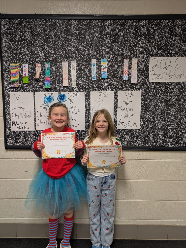 A boy and girl hold up their award certificates while standing in front of a bulletin board decorated with the winning bookmarks. 