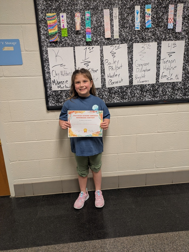 A girl holds her award certificate while standing in front of a bulletin board decorated with the winning bookmarks. 