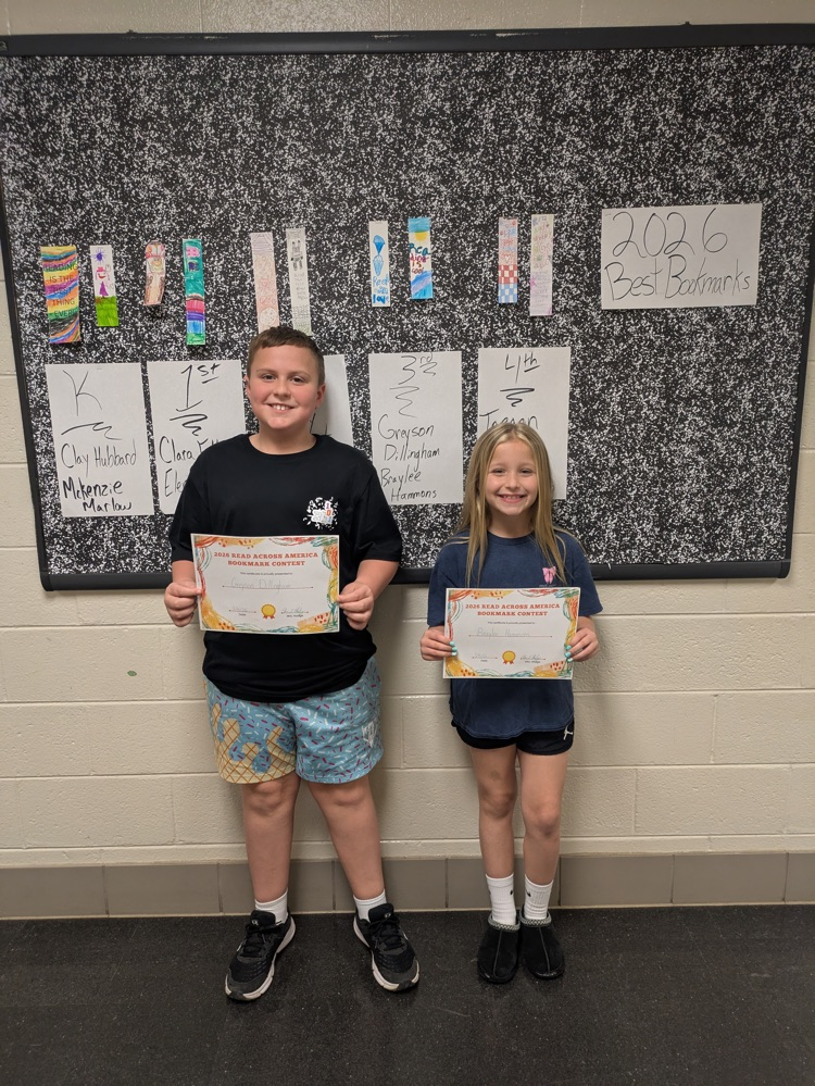 A boy and girl hold up their award certificates while standing in front of a bulletin board decorated with the winning bookmarks. 
