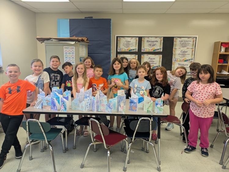 A class of students stands behind a classroom table displaying their paper media perspective art projects  