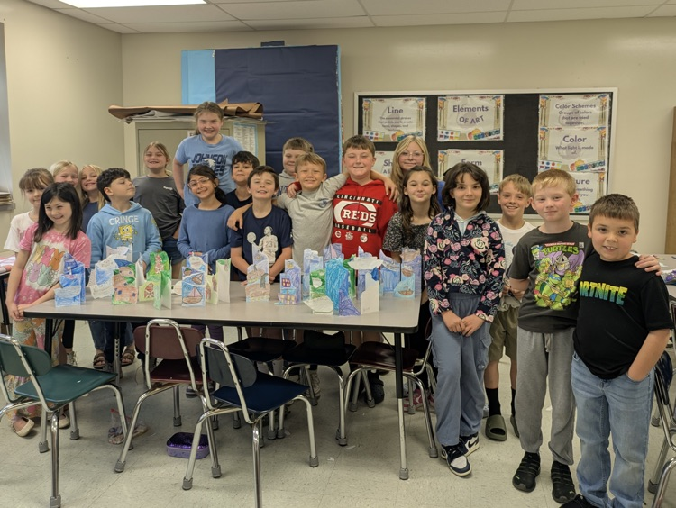 A class of students stands behind a classroom table displaying their paper media perspective art projects. 