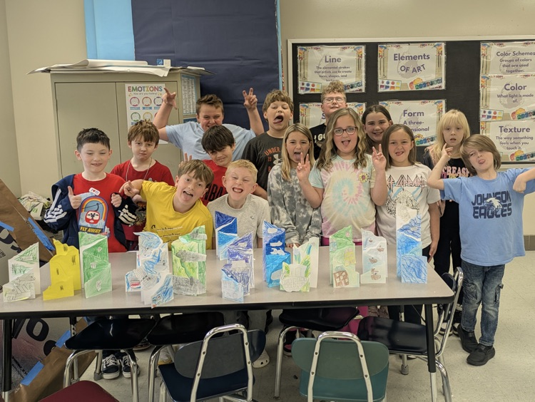 A class of students stands behind a classroom table displaying their paper media perspective art projects. 