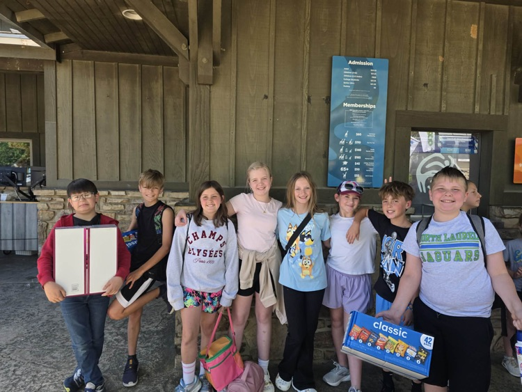 A group of fifth graders smiles for the camera as they gather lunch supplies  