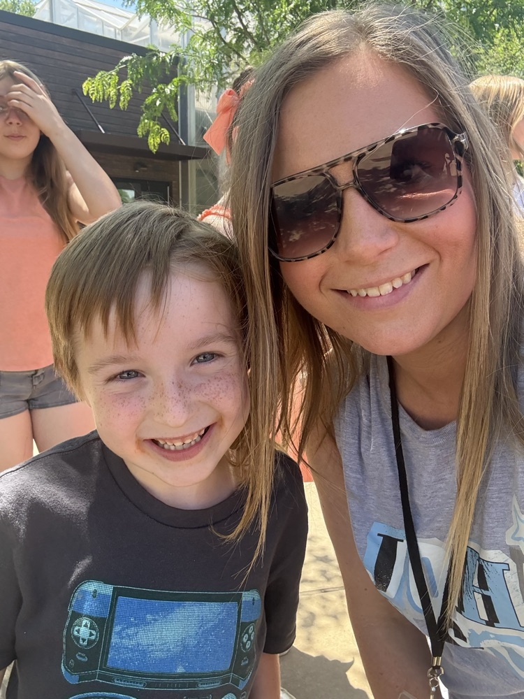 A kindergarten boy leans toward his class’s assistant as they smile for a selfie  