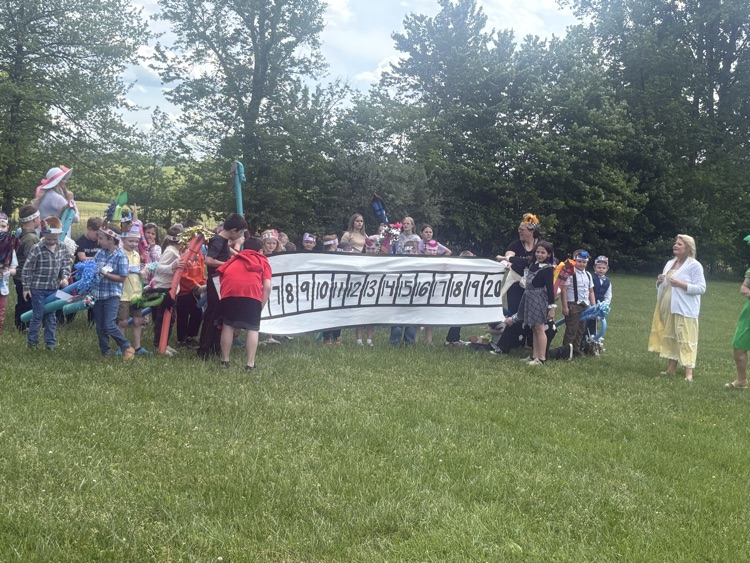 A class of children holding stick horses lineup in a field behind a banner made to resemble a horse race starting gate.