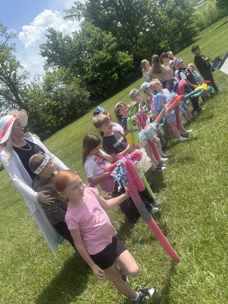 A group of students stand in line in front of their teacher waiting to start a race. They hold pool noodle stick horses.