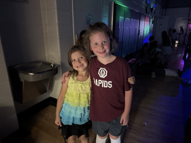 Two girls stand shoulder to shoulder, smiling for a picture in the school gym, which is darkened for the school dance  