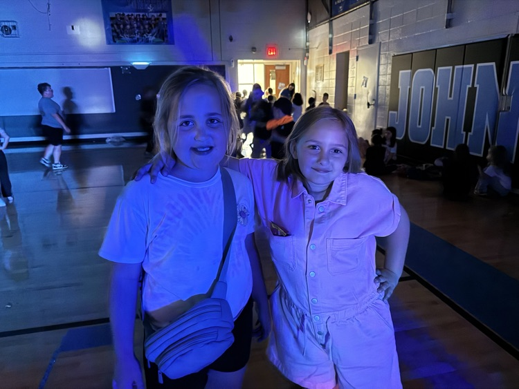 Two girls stand shoulder to shoulder, smiling for a picture in the school gym, which is darkened for the school dance. 