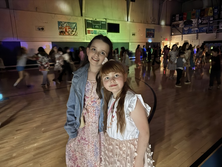Two girls stand shoulder to shoulder, smiling for a picture in the school gym, which is darkened for the school dance. 