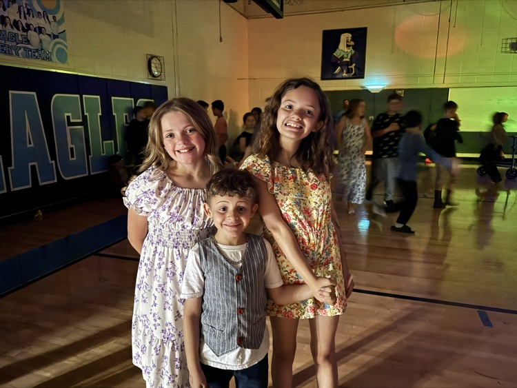 Two girls and a younger boy stand together in the school gym, which is darkened for the school dance. 