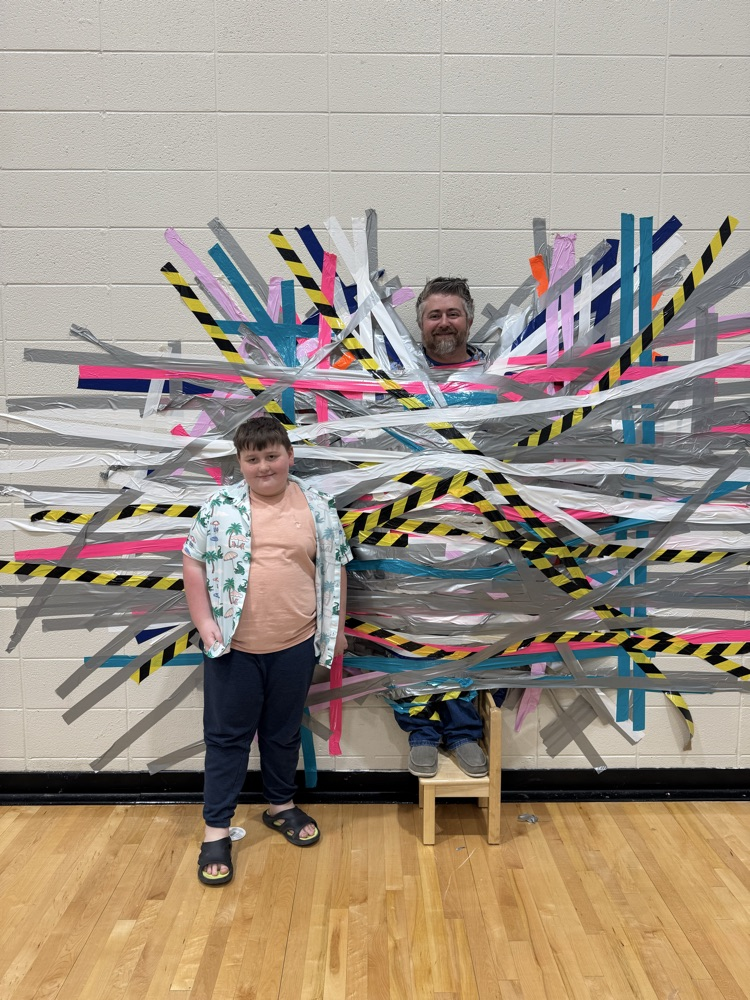 A man stands on a small chair, with large amounts of multicolored duct tape holding him to the wall. A boy stands to his left and smiles for his picture