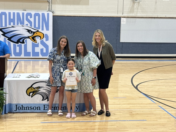 Featured left to right, Mrs. Ledington (in a blue floral dress), a female student holding her awards, Mrs. Maggard (in a green dress), and Mrs. Bailey (in a black dress with green sweater) stand in the gym in front of the awards table