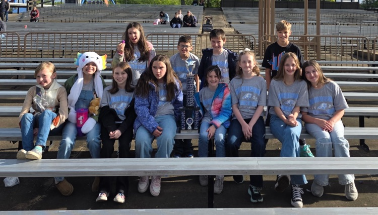 A group of students wearing matching gray choir t-shirts sit in three rows on a set of bleachers