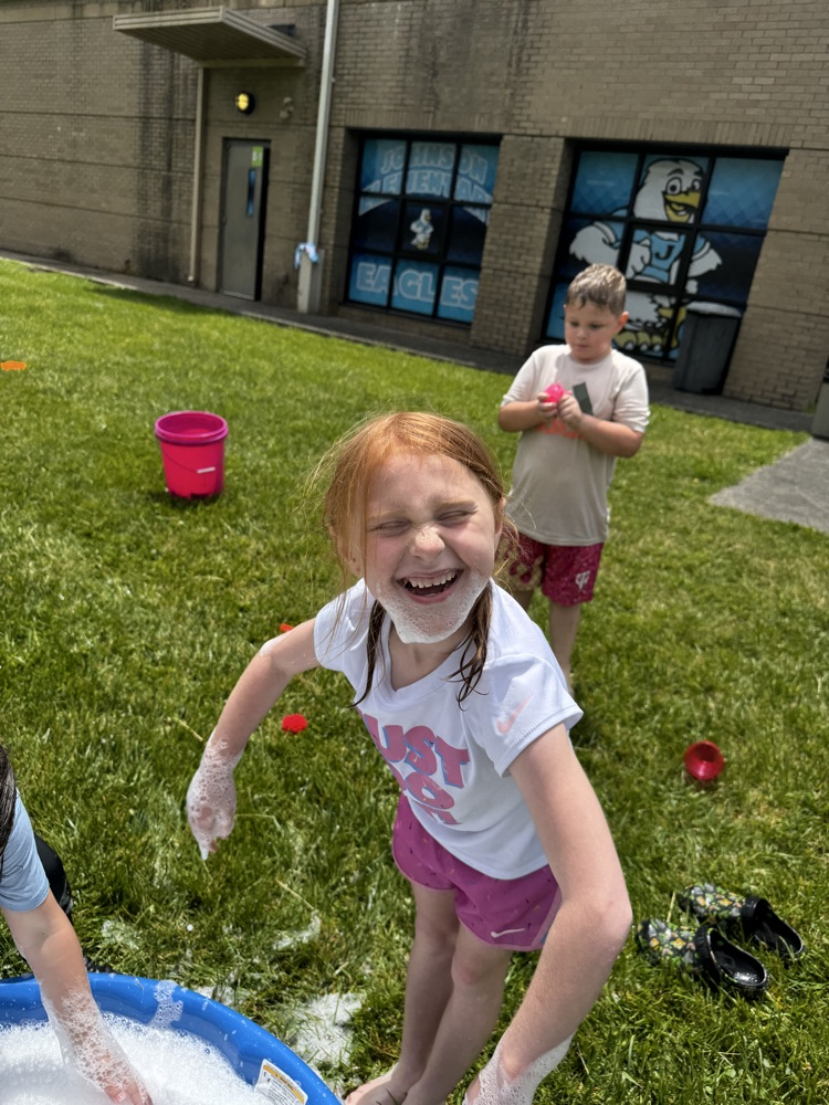 A girl in a white t-shirt and pink shorts plays in a kiddie pool filled with bubbles. she has bubbles on the bottom of her face. A boy stands behind her and is playing in the water as well.