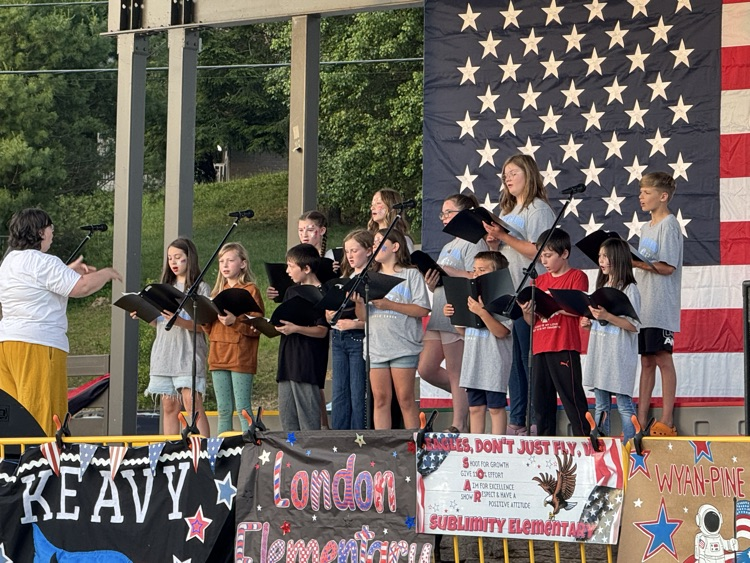 A choir and their director perform on a stage decorated with patriotic banners. They hold black music binders