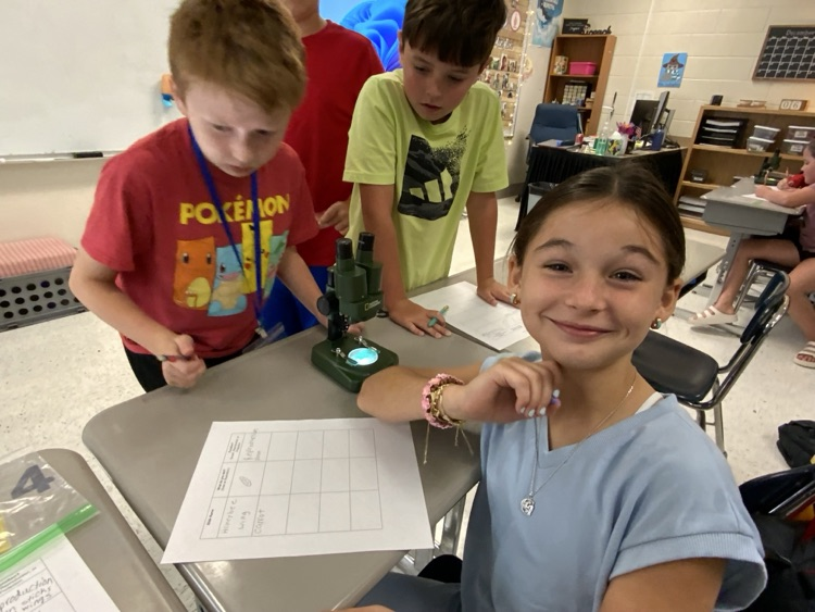 Students gather around a microscope sitting on a classroom desk. They are taking notes about their observations, and one girl in a blue shirt looks up and smiles at the camera  