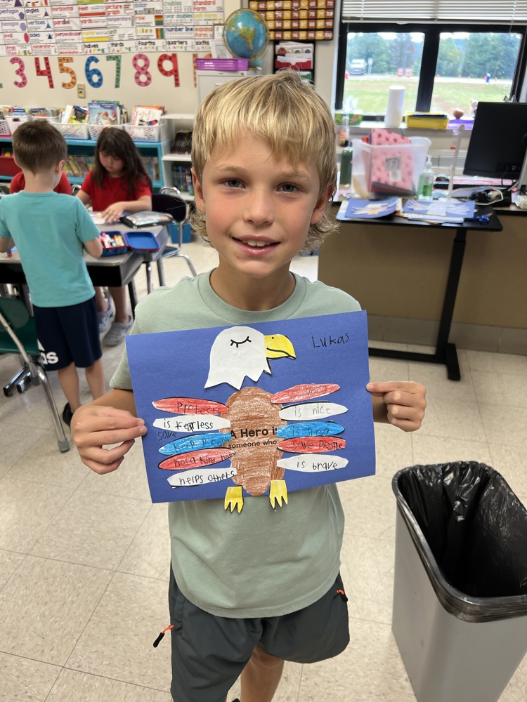 A boy smiles while holding his red, white, and blue eagle project highlighting the characteristics of a hero. 