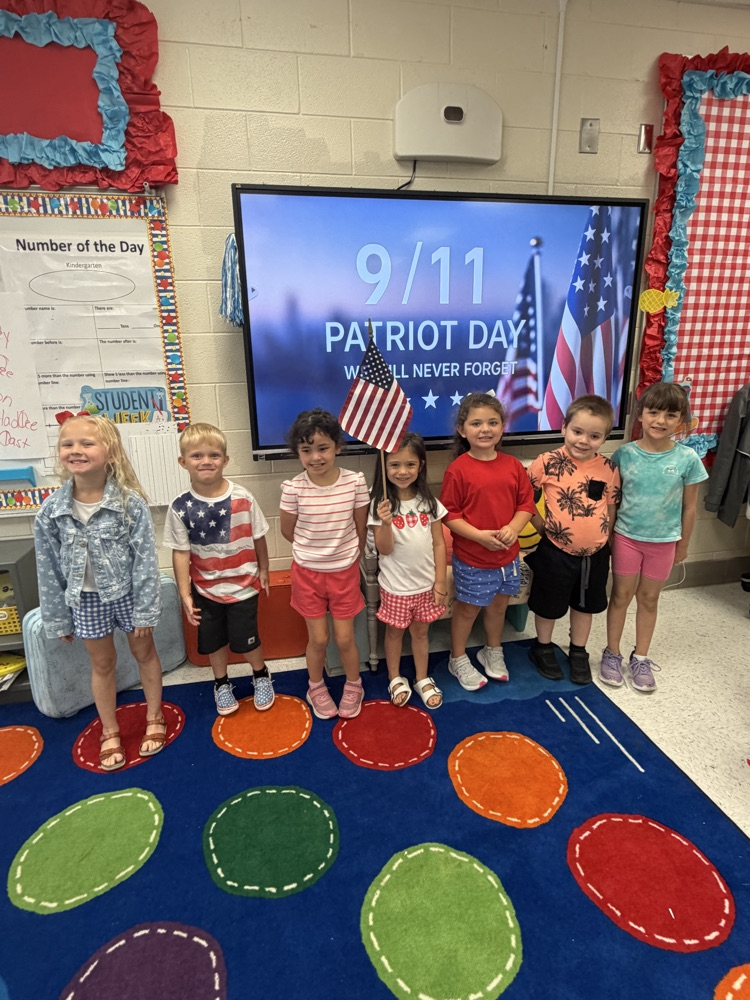A line of seven kindergarteners stand in front of a screen with patriotic imagery. The child in the middle holds up an American flag  