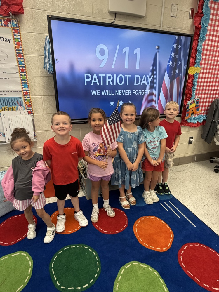A group of six children stand in a line in front of a screen displaying patriotic imagery. The middle child holds an American flag  