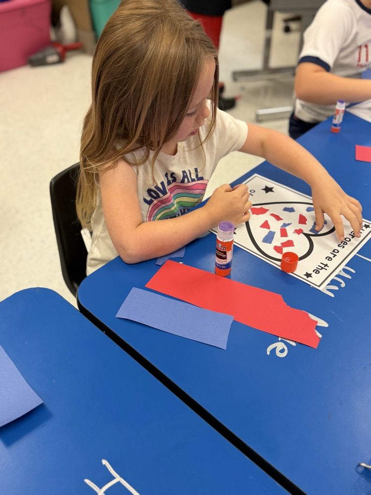 A girl sits at a blue table working on her torn paper heart art project  