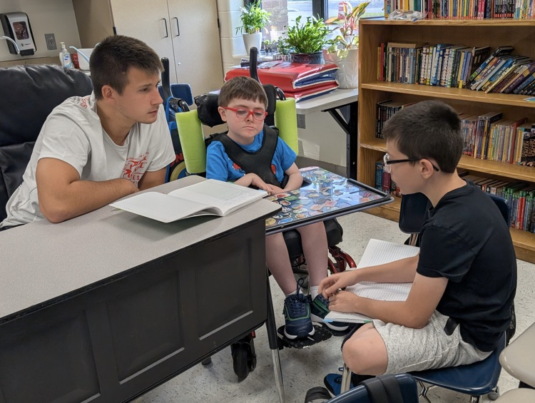 A male instructional assistant sits with two boys, one in a wheelchair, while they discuss their assignment