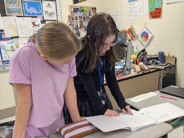 two girls, one in a purple shirt, and one in black, look down on their notebook together.