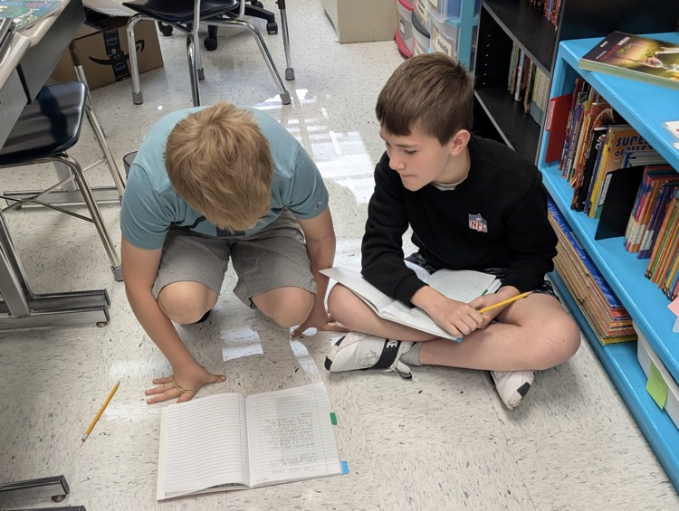 two boys sit in a classroom floor working cooperatively in one notebook.