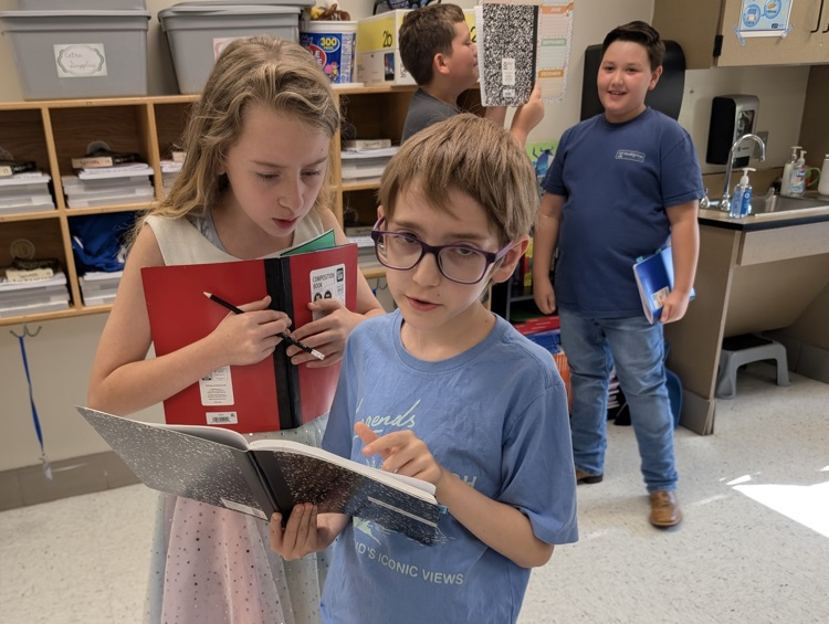 A boy and girl look into a notebook together standing in the middle of a classroom. Behind them two boys are also working collaboratively.