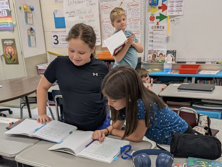 two girls staying together in front of their desks, each working in their notebooks. One leans forward onto the desk.