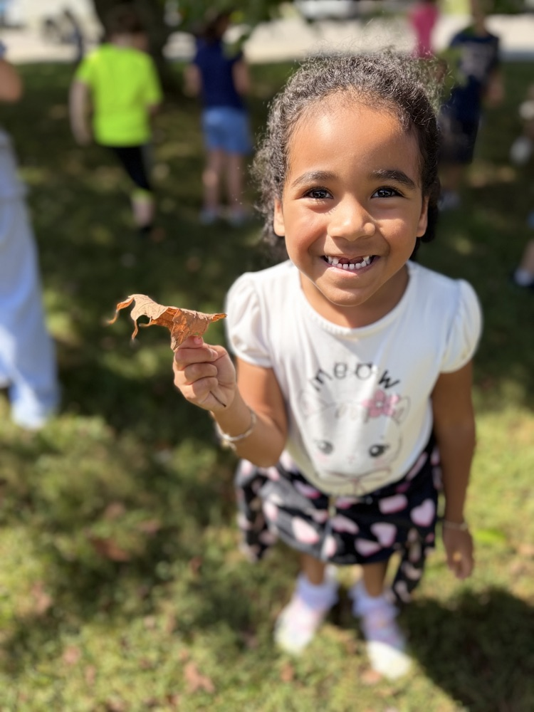 A young girl in a white t-shirt stands outside in the grass. she holds up a brown leaf