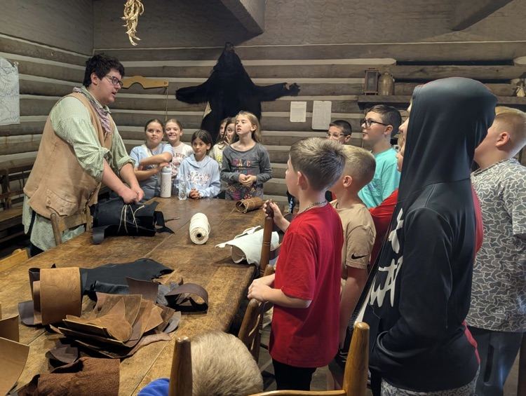 A man in period clothing stands at a table, covered with leather samples of various colors. Students stand around him and listen to his demonstration.
