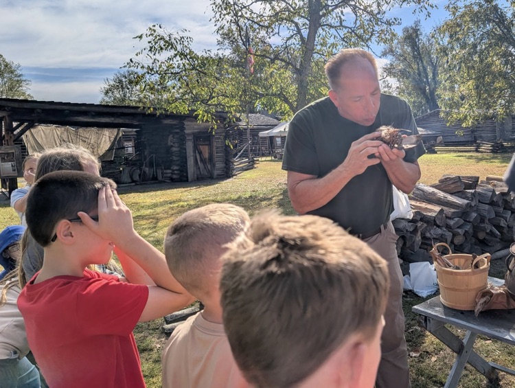 A man demonstrates using flint to make a fire to a small group of boys. He blows on his kindling to start the fire.