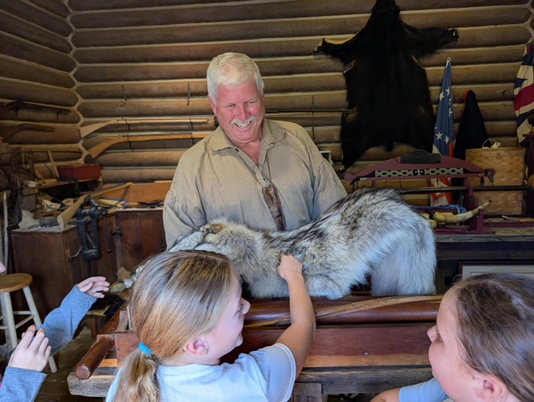 A man in Pioneer style clothing holds a wolf pelt. A girl touches the pelt while two other students look on.