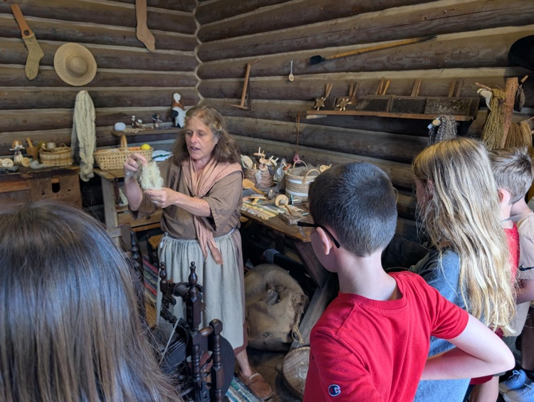 A woman in pioneer style clothing holds up wool that she is using to make thread. She stands in front of a spinning wheel. Students stand up in the foreground of the picture watching her.