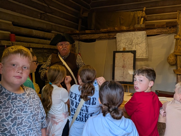 A man and a tricorn hat and period clothing stands in front of a small group of children who listen to his talk about surveying. Behind him a table is strewn with maps.