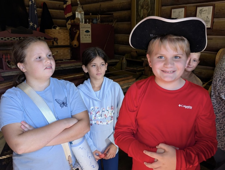 two girls in blue shirts, look to the right where a boy in a red, long sleeved T-shirt smiles while wearing a tricorn hat.