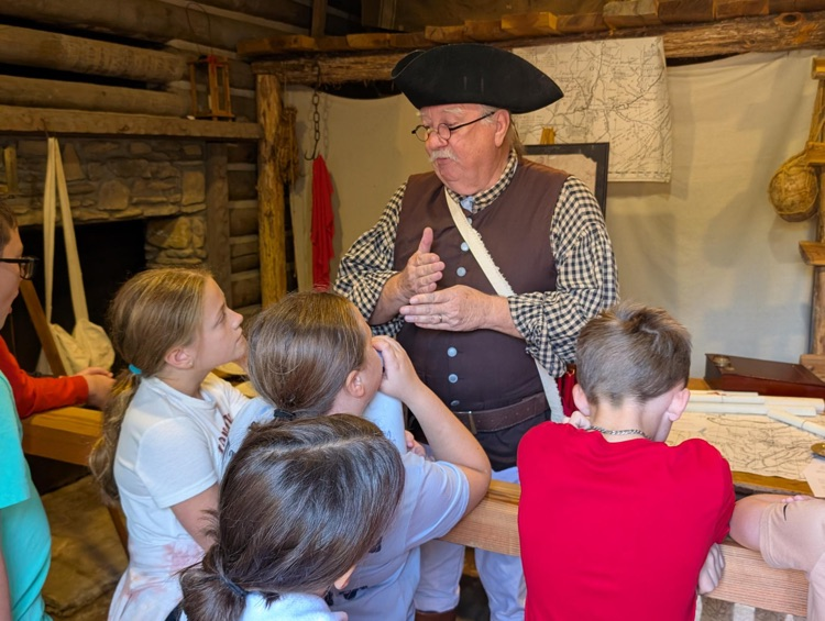 A man and a tricorn hat and period clothing stands in front of a small group of children who listen to his talk about surveying. Behind him a table is strewn with maps.