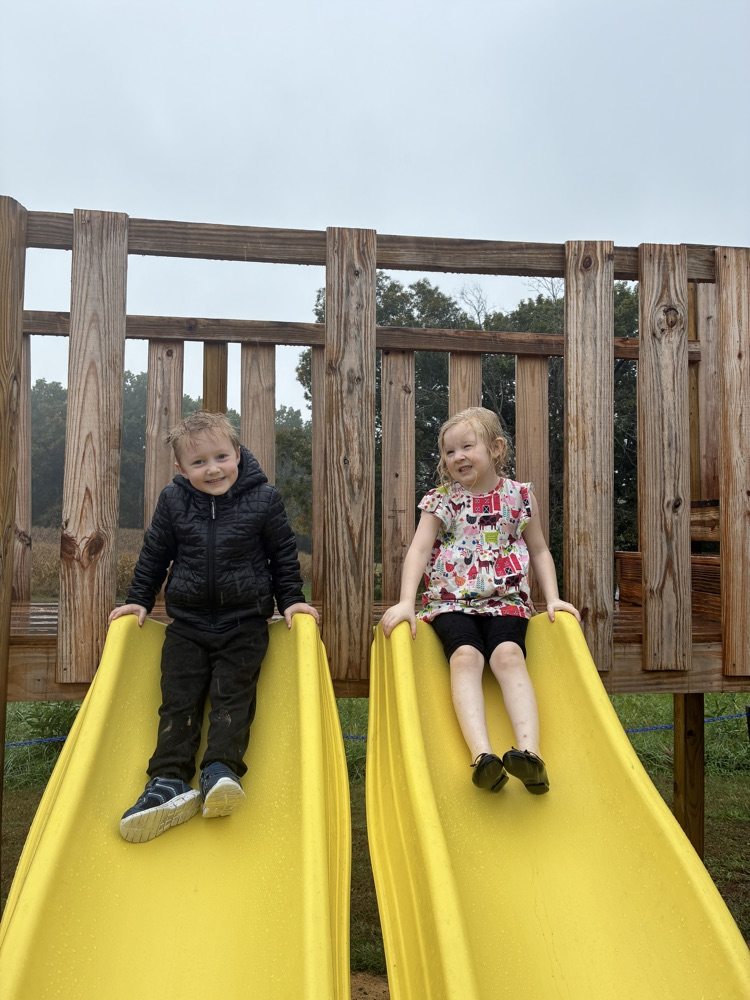 A boy in black and a girl in black white and pink dress sit on tandem yellow slide slides.