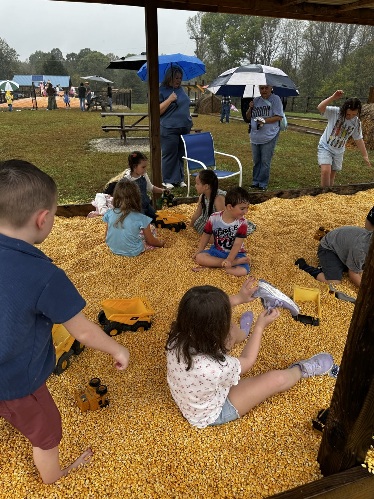 A group of children sit together in the corn bin playing with trucks.