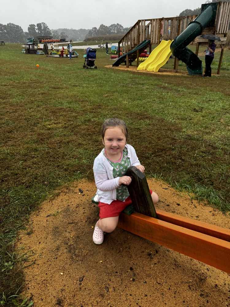 A girl in a white sweater, green shirt and red shorts rides a seesaw.