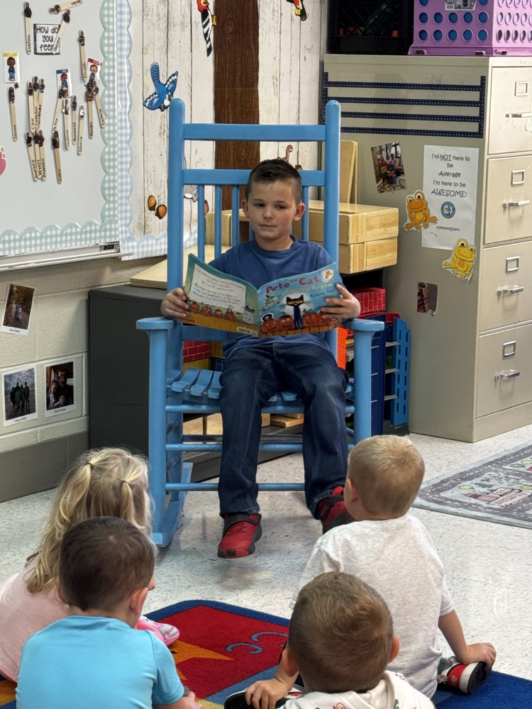 A boy in a blue T-shirt and jeans sits in a blue rocking chair. He holds a Pete the cat book in his hands. In front of him, four students are seen sitting on a rug, facing him and listening to him read.