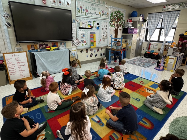 A wide shot of the Johnson preschool classroom shows a boy sitting in a blue rocking chair, reading a book to a classroom of preschool students. The students sit on a brightly colored rug with their backs to the camera facing the boy.
