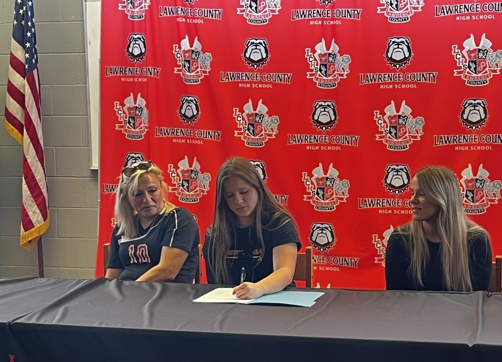 Three women seated behind a table, one signing a paper. Behind them, a red backdrop with text and logos, and a flag on the left.