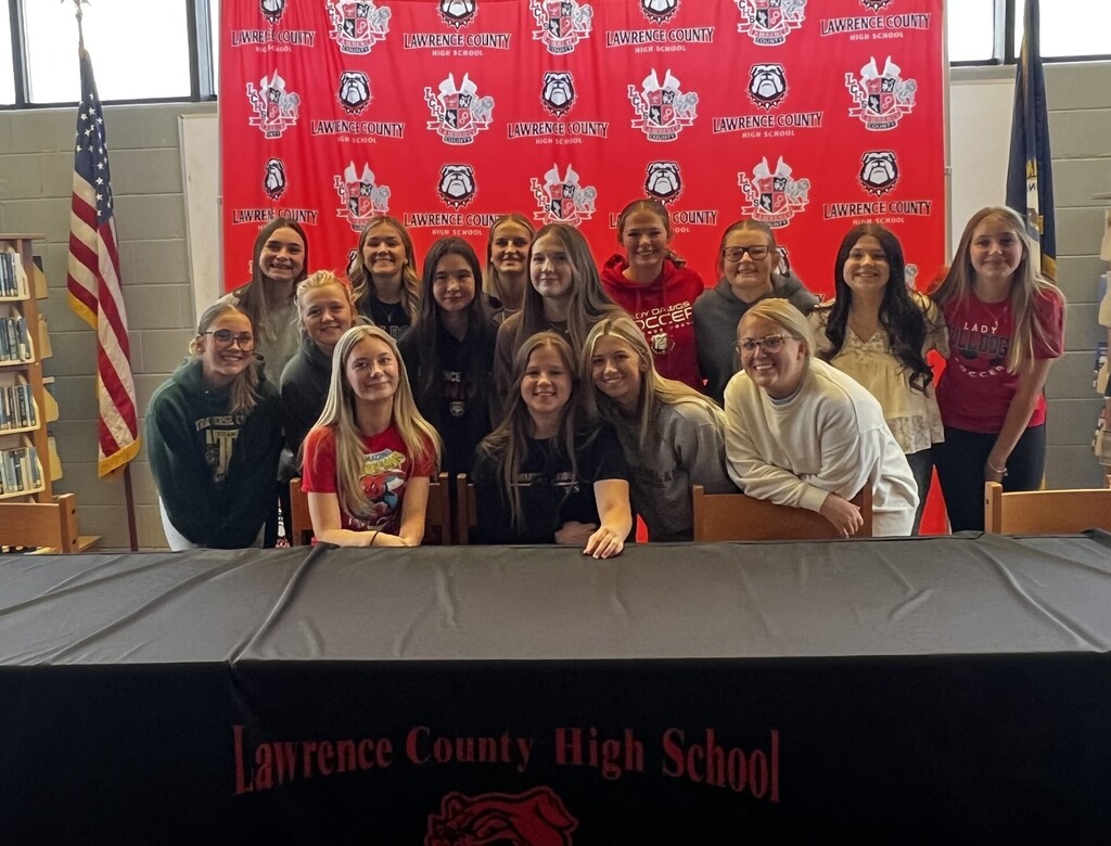 A group of young women stand behind a table with a banner reading 'Lawrence County High School'. Some wear team colors.
