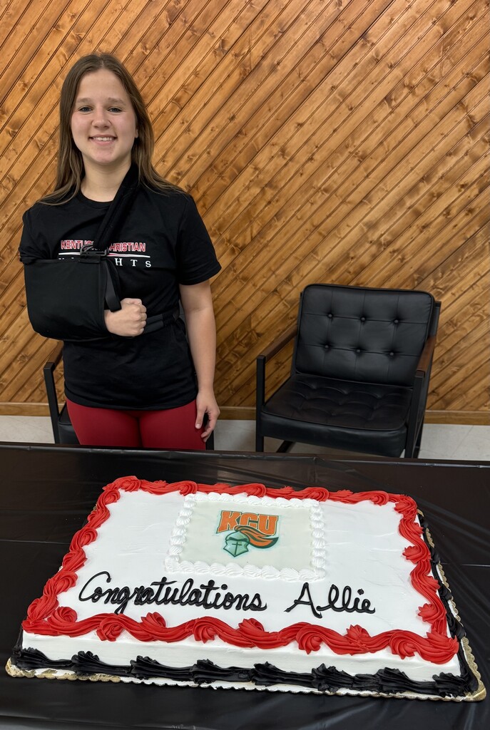 A woman with an arm sling stands near a wooden wall, beside a cake on a table. The cake reads "Congratulations Allie".