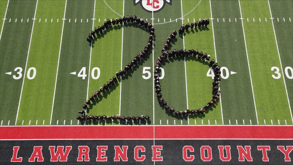 Aerial view of a football field with a group forming the number 25. The field has yard lines and is marked with red and black lines.