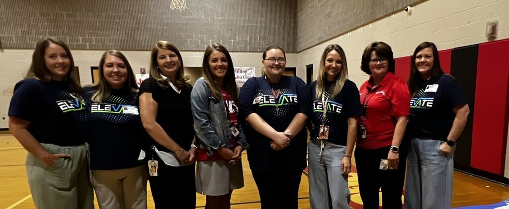 Six women in various tops and pants, possibly in a gym or sports hall, with two in red shirts.